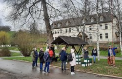 Teilnehmergruppe bei einer erlebnispädagogischen Kletteraktion mit Getränkekisten im Freien, Kinder und Erwachsene schauen zu, Hintergrund Gebäude und Bäume.