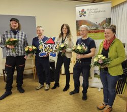 Fünf Personen mit Blumensträußen posieren vor Verbands-Roll-ups.